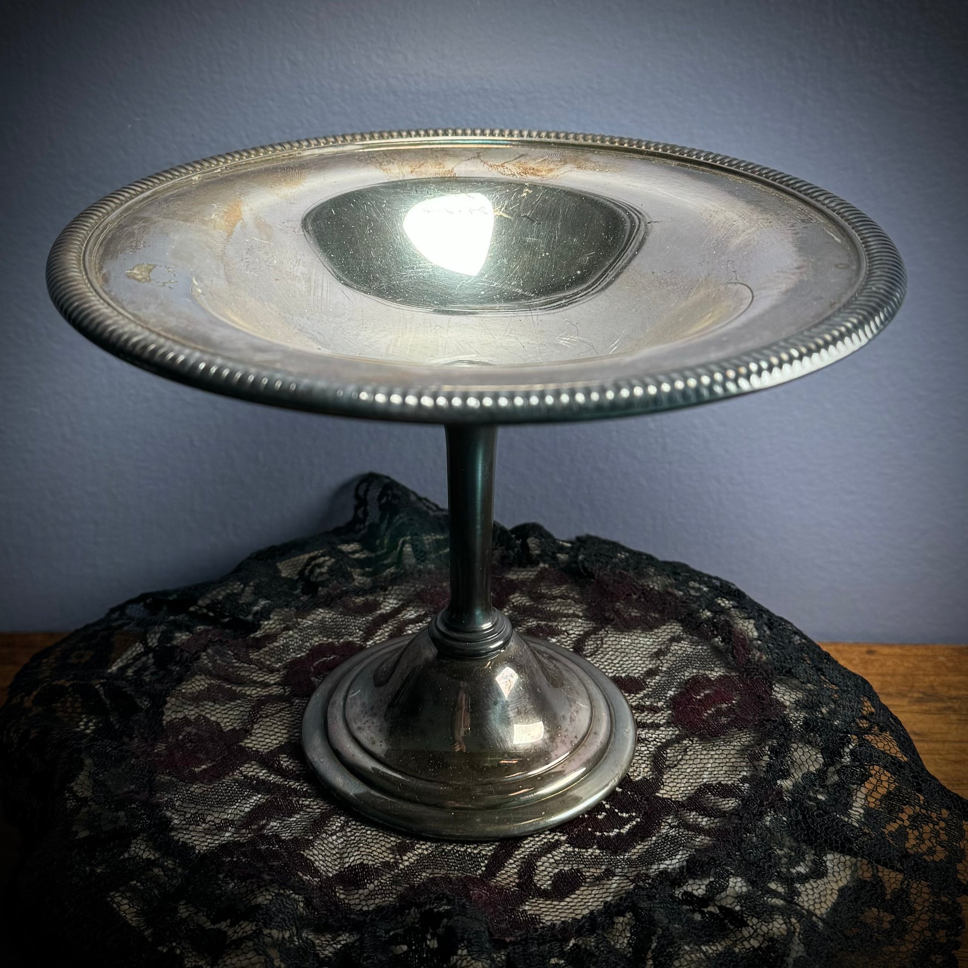 A vintage silver plate bowl with a step and round base sits on a black doily