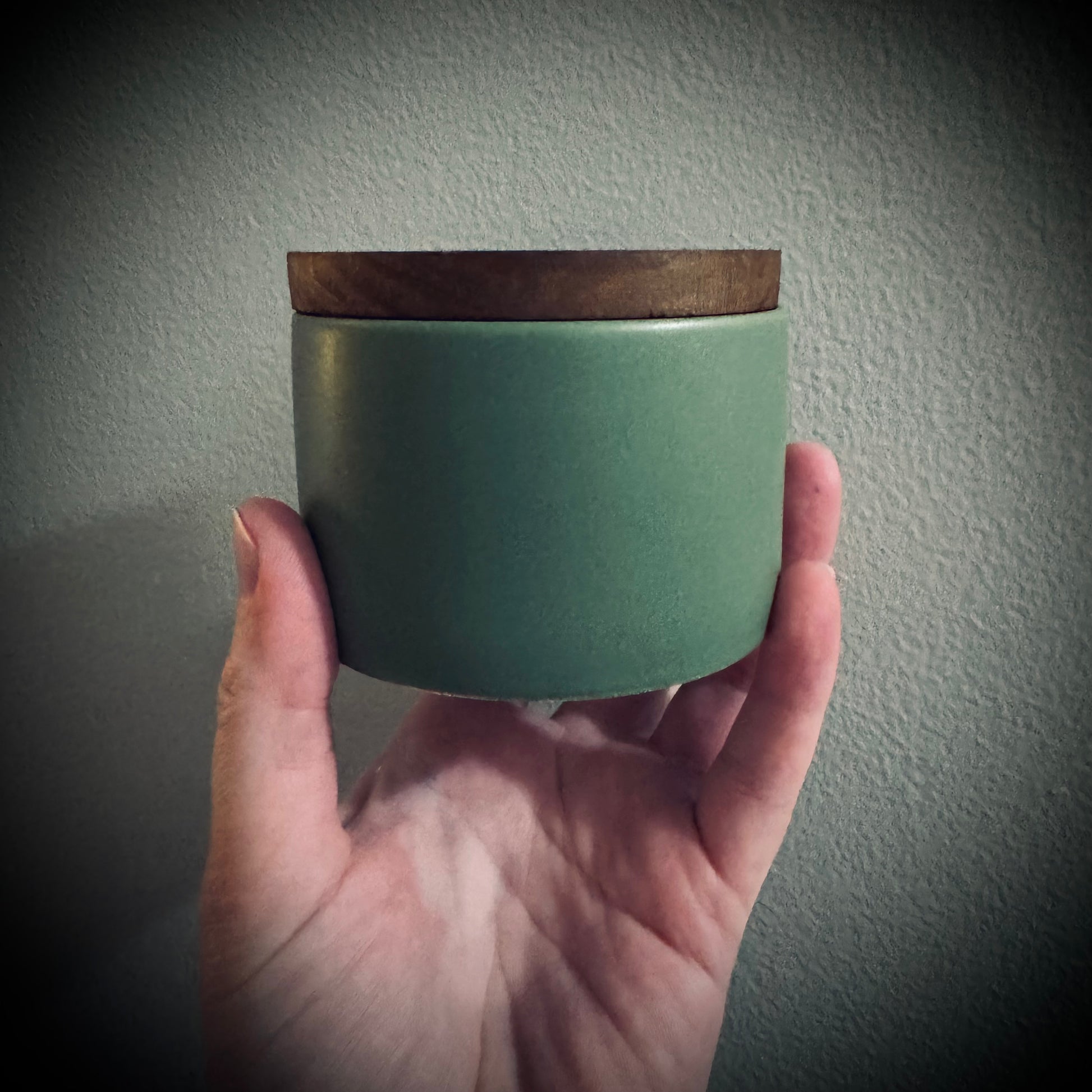 Hand holding a green ceramic candle jar with a wooden lid.
