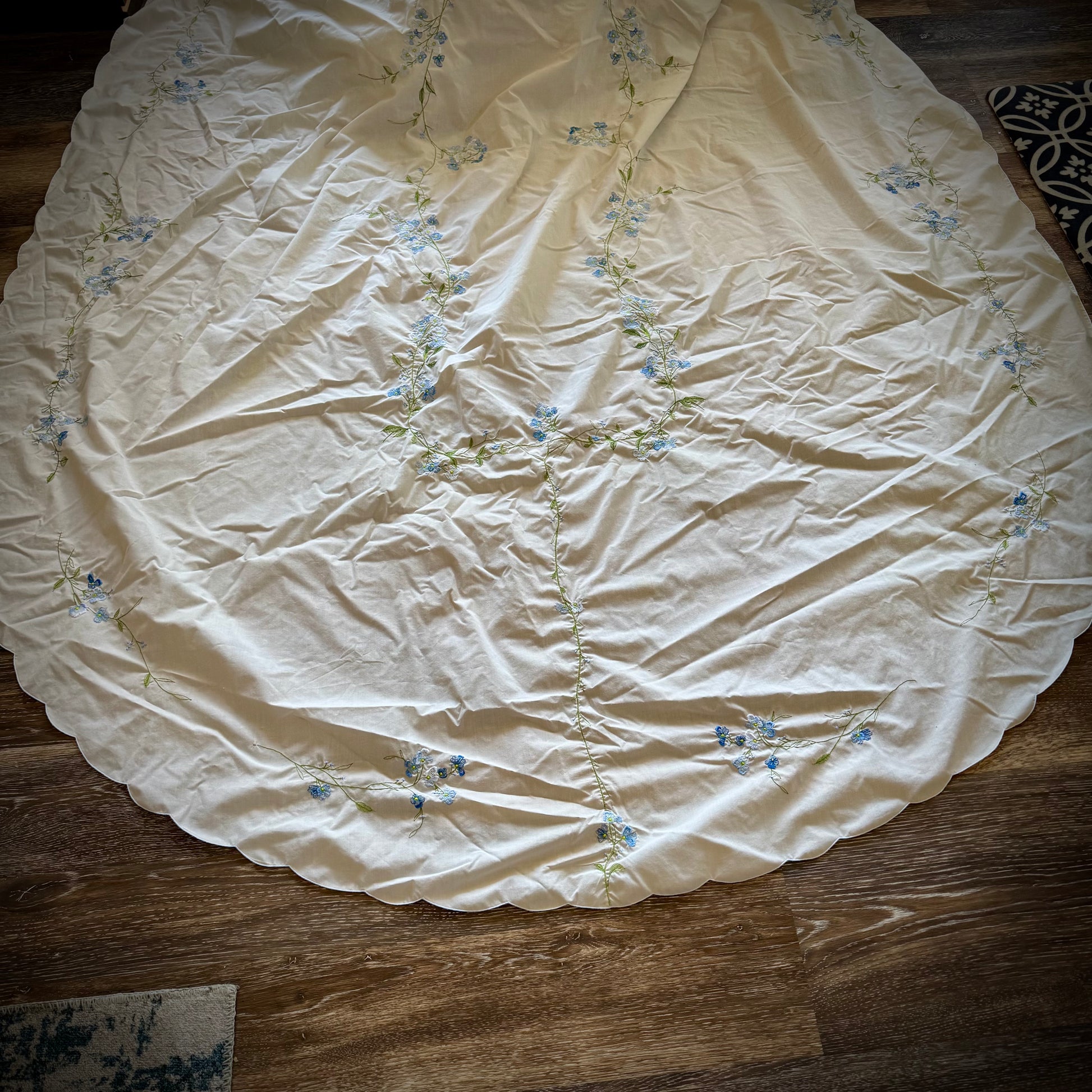 Vintage embroidered oval tablecloth laid out on a surface, primarily white with blue floral embroidery, showing signs of age-related discoloration.