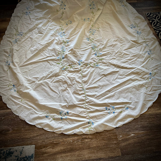 Vintage embroidered oval tablecloth laid out on a surface, primarily white with blue floral embroidery, showing signs of age-related discoloration.