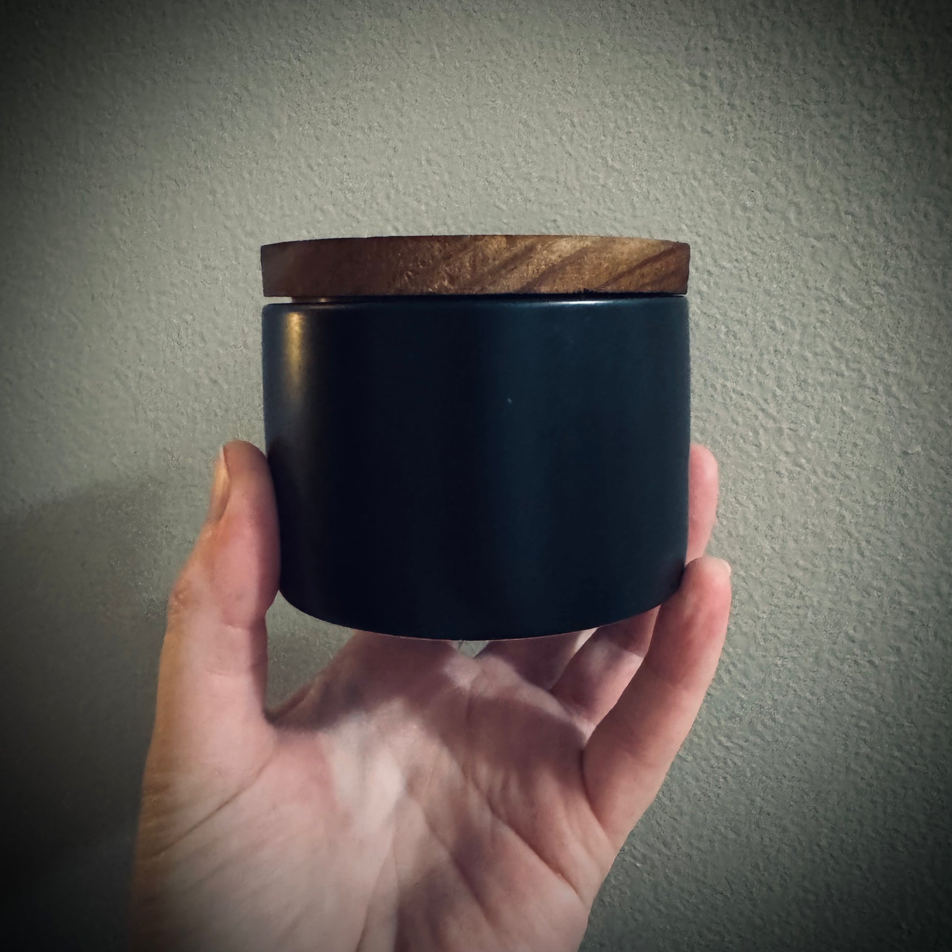 Hand holding a black ceramic candle jar with a wooden lid.