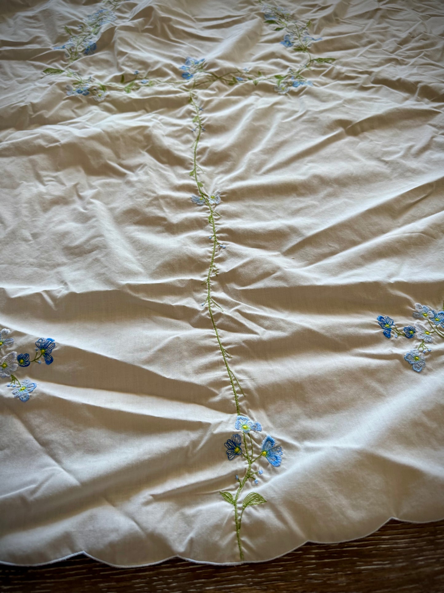 Close up of a vintage embroidered oval tablecloth laid out on a surface, primarily white with blue floral embroidery, showing signs of age-related discoloration.