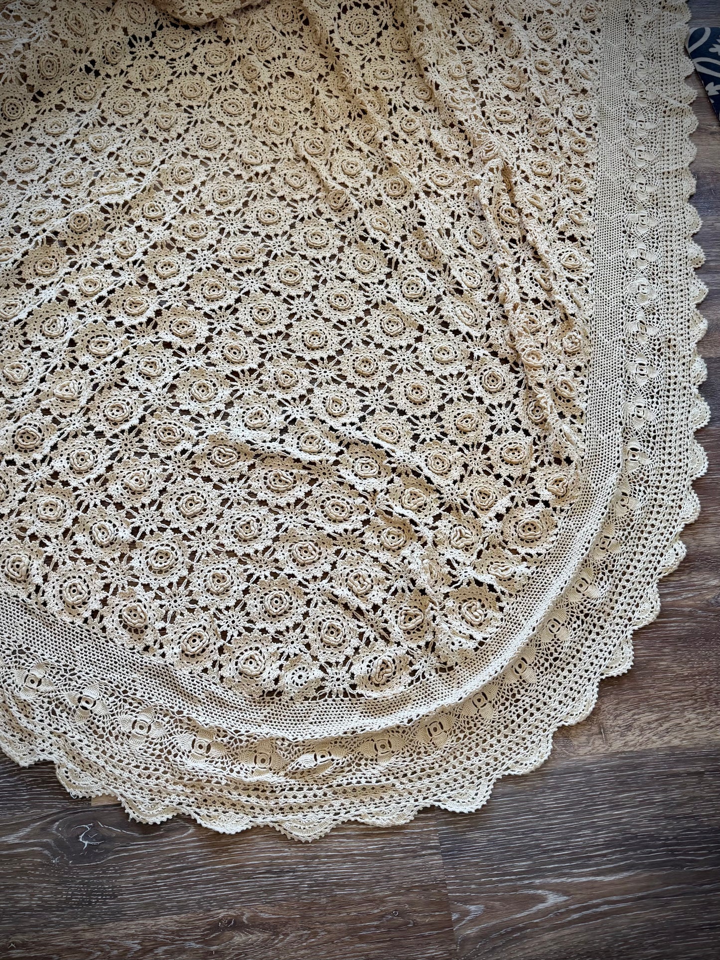 A beige crochet coverlet featuring a floral rosette pattern and a rounded scalloped edge.