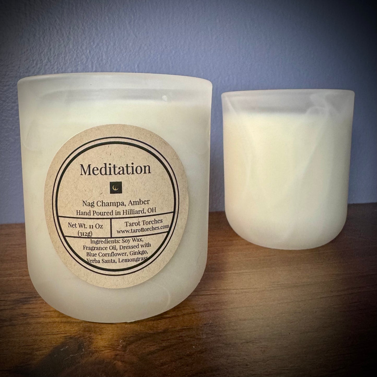 A white candle with the label "Meditation" sits on a wooden surface in front of an unlabeled white candle.