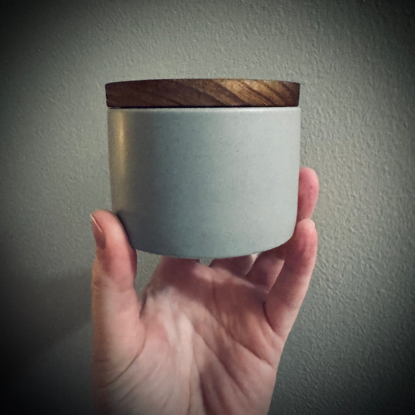 Hand holding a gray ceramic candle jar with a wooden lid.