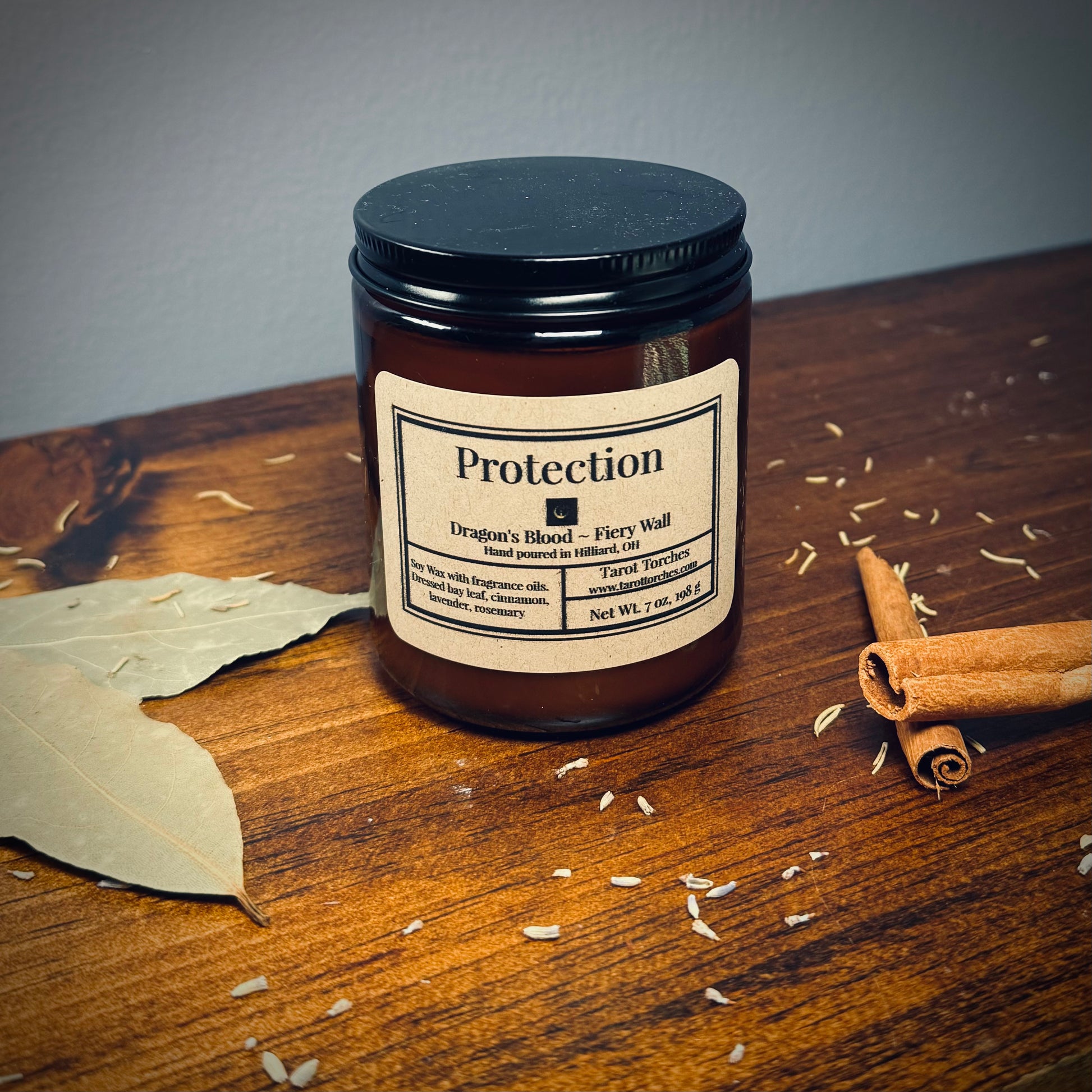 An amber glass candle with a black lid and a label that reads "Protection" on a wooden surface with scattered herbs.