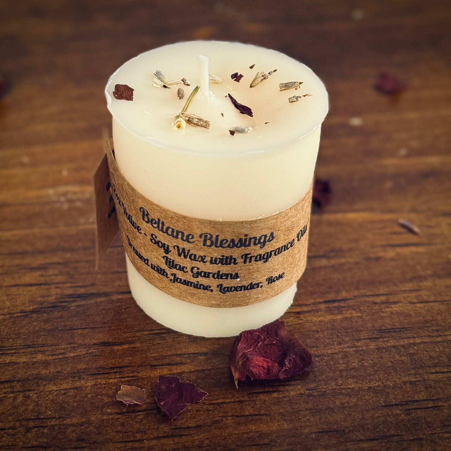 A white votive candle with a label reading "Beltane Blessings" sits on a wood surface surrounded by herbs.