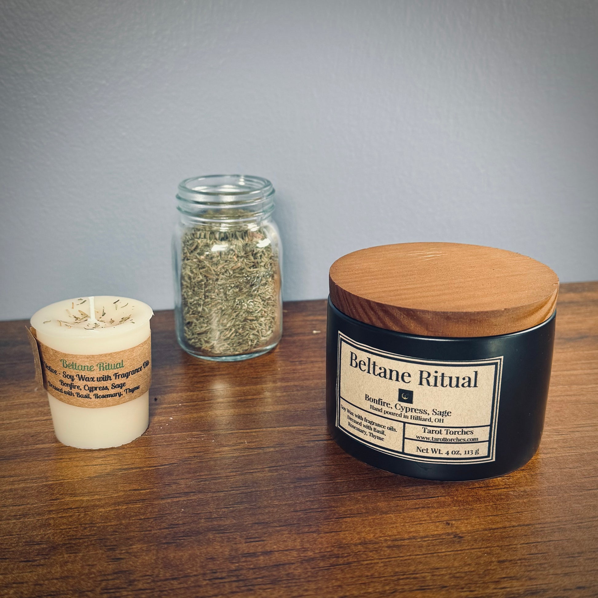 A white votive and a black ceramic candle both labeled "Beltane Ritual" sit on a wooden surface with a glass jar of herbs in between.