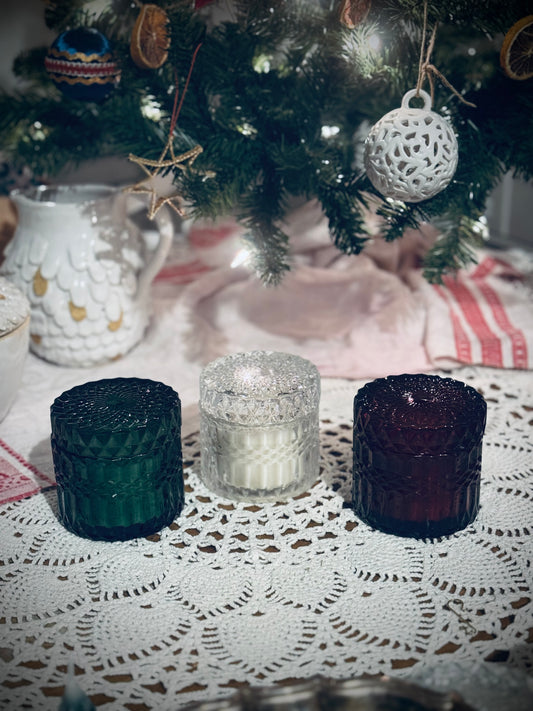 Three glass candles in green, red, and clear colors, placed on a lace tablecloth in front of a decorated Christmas tree.