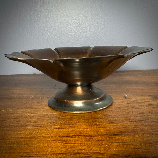 A vintage ritual bowl with a scalloped edge and a deep dark copper color, placed on a short and sturdy stand.