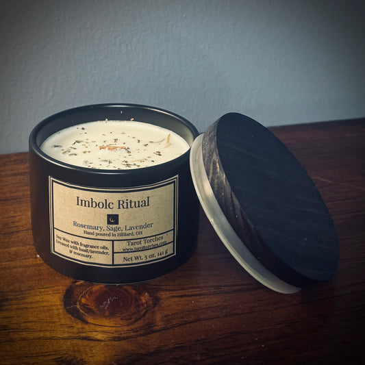 A black ceramic candle with a lid, labeled 'Imbolc Ritual' with notes of rosemary, sage, and lavender, sitting on a wooden surface.