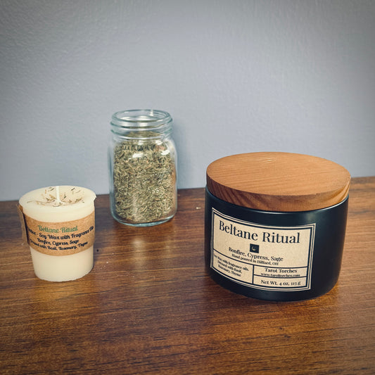 A white votive and a black ceramic candle both labeled "Beltane Ritual" sit on a wooden surface with a glass jar of herbs in between.